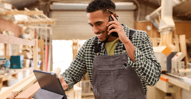 Male Apprentice With Digital Tablet Working As Carpenter In Furniture Workshop Making Phone Call