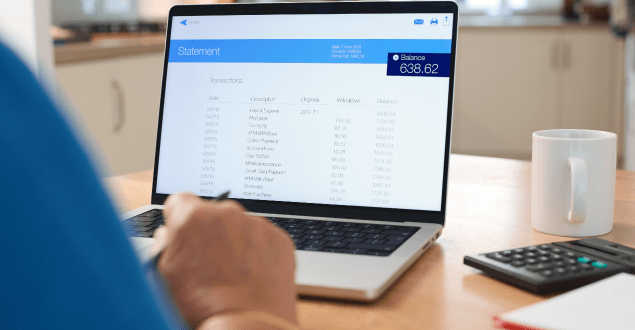 A person sits at a kitchen table, focused on reviewing their bank statement displayed on a laptop screen.
