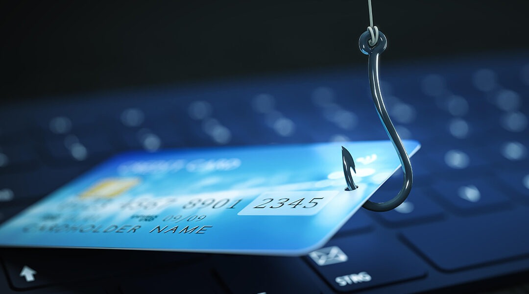 Blue credit card on a laptop keyboard being pierced by a fishing hook, illustrating phishing attacks online.